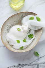 Beige bowl with burrata cheese in brine and green basil leaves, vertical shot on a white granite background, high angle view