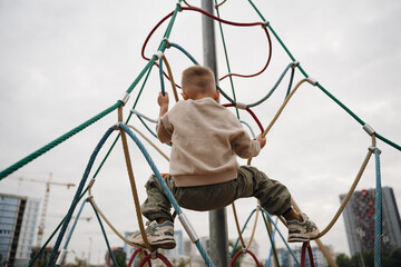 On a summer day, a boy climbs an alpine grid in a park on a playground. Playground in a public...