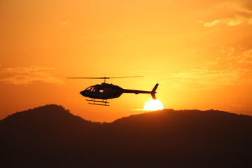 The silhouette of a helicopter Bell 206, sharply defined against a vibrant orange sunset. The sun, sits low on the horizon behind the aircraft, casting long shadows on the distant, dark hills. © Davison