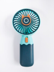 A small portable handheld fan in turquoise and dark blue color with an orange button, placed on a white background © SEPRIA