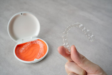 Clear dental aligner in hand and orange case on table. Dental orthodontic mouthpiece.