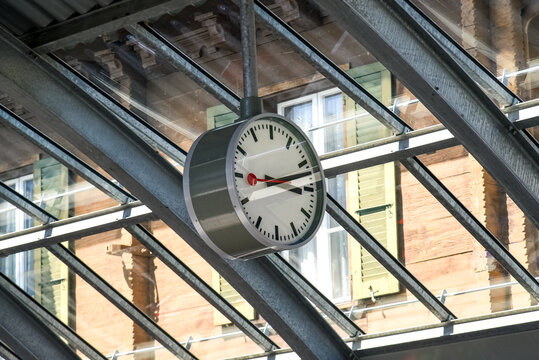 Swiss railway station clock with its distinctive minimalist design and red second hand, hanging under a glass roof structure. A symbol of precision and punctuality in Swiss train stations