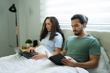 Couple relaxing in bed: woman using laptop, man reading book
