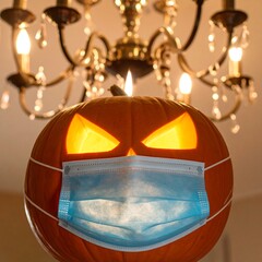 A jack-o'-lantern pumpkin, wearing a face mask, hangs beneath a chandelier