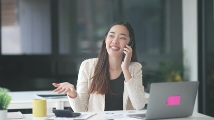 Professional Asian businesswoman working at office desk, communicating on phone with laptop and calculator, expressing cheerful confidence during business interaction - Powered by Adobe