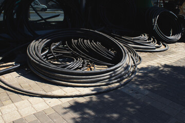 A large pile of coiled black industrial hoses rests on a brick pavement, with a few strands...