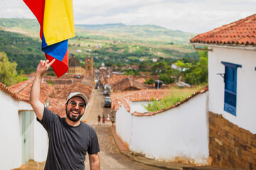 Smiling young Latino man pointing at the Colombian flag in the colonial town of Barichara, Colombia, expressing national pride, culture, heritage tourism, joy and authentic travel adventure.