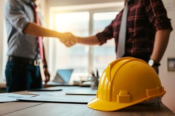 Two professionals shaking hands in a bright office, symbolizing partnership and project success with safety gear nearby.