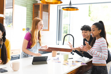 Teen friends in kitchen laughing and chatting with tablet and coffee mugs