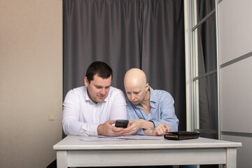 Worried couple sitting at home, checking bills and financial documents while using a smartphone, woman with bald head from cancer treatment and man in white shirt focused on screen