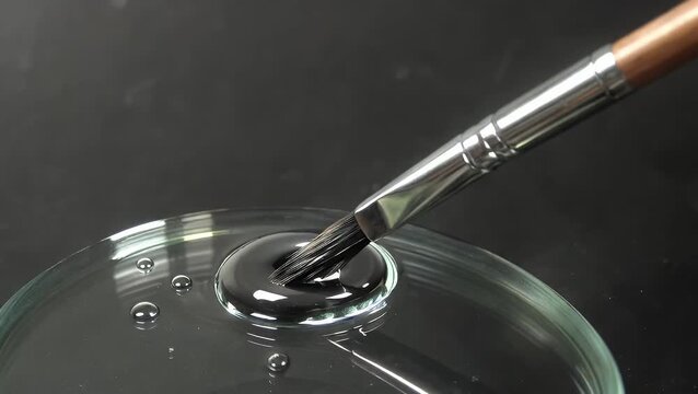 macro photographic capture depicting metallic-finished artist's brush being drawn through liquid medium adjacent to transparent surface, creating abstract pattern with dark contextual setting