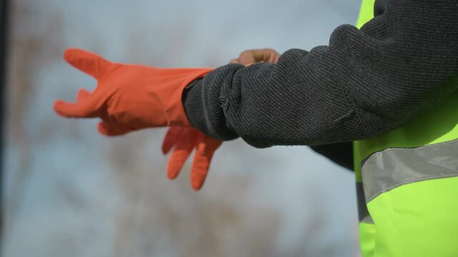 Close-up of worker wearing dark sweater and high visibility vest putting on bright orange protective gloves, preparing for safety tasks in outdoor construction
