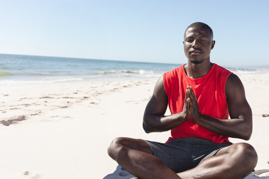 Meditating on beach, African American man in red shirt enjoying peaceful moment, copy space