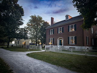 Historic brick building at dusk with glowing lights in windows, white fence, and tree-lined path at...