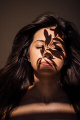 Young woman with long dark hair, eyes closed, illuminated by soft light, with intricate shadows cast on her face, creating a serene and contemplative atmosphere in a minimalist setting