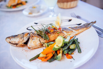 Grilled fish served on a white plate with colorful vegetables, garnished with herbs, showcasing a delicious seafood dish in a bright dining setting