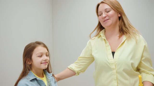 Close up of caring mother in yellow shirt kissing daughter on forehead after cleaning together with broom and dustpan, showing love and warmth in bright laundry space with tiled floor