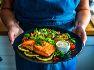 Woman holding plate with fried salmon steak with boiled broccoli and mini carrots, fried potatoes and mayonnaise, close-up