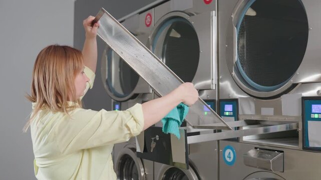 Manager returns cleaned lint filter into drawer of industrial washer, guiding mesh panel with turquoise towel, routine maintenance inside laundromat, stainless units lined in row, hygiene focus