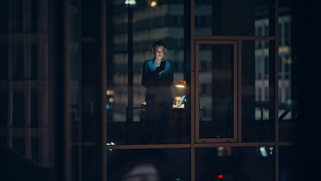 Silhouetted businesswoman standing window typing smartphone alone at night hours