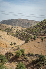 Dry terraced hills in Morocco with scattered green shrubs and winding paths cutting through the arid slopes under a pale sky