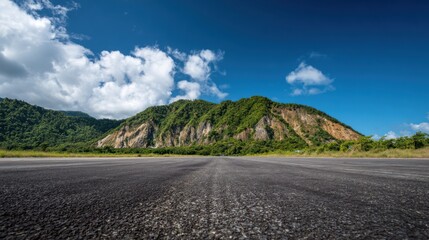 Fototapeta premium A wide asphalt road leads into the distance, framed by lush green mountains. Fluffy white clouds float in a bright blue sky, creating a picturesque, tranquil setting.