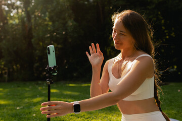 Young woman in sportswear stretching body on fitness mat at public park. Beauty blogger filming yoga in morning park. Vlogger live streaming. User-generated content UGC. Part of a series. Soft focus