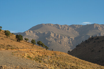 Rocky hills and rugged mountain peaks in Morocco under a clear blue sky, with dry slopes and scattered trees on the arid landscape