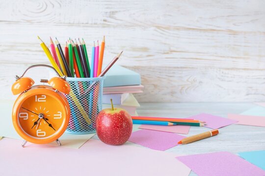 Back to school concept. Alarm clock, stack of books, apple, colored pencils and pastel colored paper on white wooden background.
