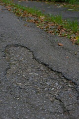 A cracked and weathered asphalt road with visible gravel and a single fallen leaf, creating a textured and moody image of urban decay, damage, and neglect.