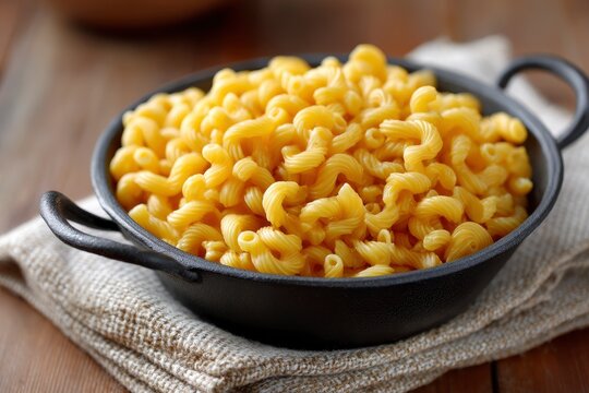 A black pan filled with spiral macaroni noodles sits on a folded beige cloth atop a wooden surface