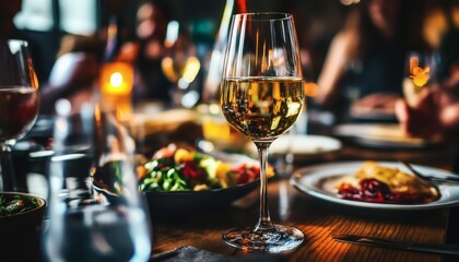 A close-up shows a glass of white wine sitting on a wooden table among plates of food, with blurred people in the background