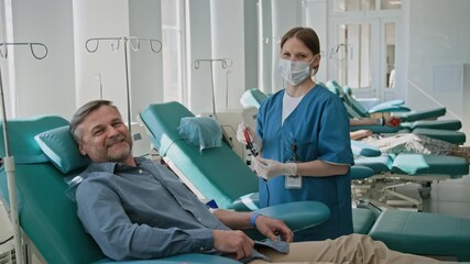 Portrait shot of masked nurse in blue scrubs and smiling middle-aged man posing for camera after donating blood in clinic - Powered by Adobe