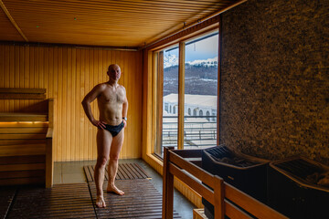 A sporty man in swimming trunks stands by the window in a sauna with a gorgeous view of the mountains.