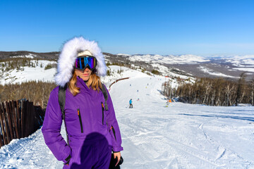 Beautiful woman in purple ski suit, hood, glasses and boots against the background of ski slope and mountains.
