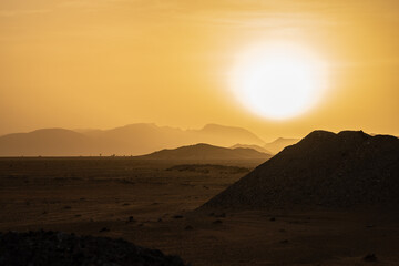 Obraz premium Golden Sahara desert sunset in Morocco with bright sun over distant mountain silhouettes and dark sand dunes creating dramatic evening landscape view