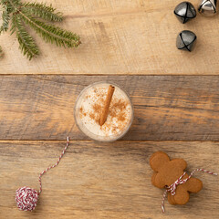 Christmas holiday traditional eggnog on wooden table with gingerbread cookies, cinnamon stick, jingle bells, pine branch, and red and white bakers twine.