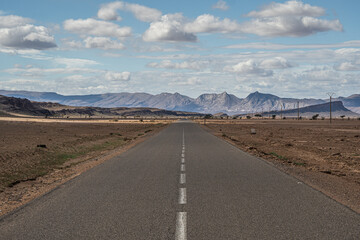 Straight asphalt road in Morocco desert stretching into distance with barren plains and rugged Atlas Mountains under cloudy blue sky panoramic view