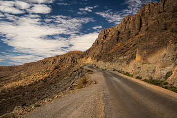 Winding mountain road in Morocco with towering rocky cliffs and a dramatic blue sky filled with white clouds on a sunny day