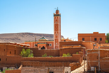 Traditional Moroccan village mosque with tall red minaret and adobe houses set against dry desert...