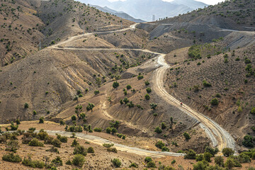 Winding dirt tracks across Morocco Atlas Mountains hills with dry rocky slopes scattered green bushes and rugged desert landscape under cloudy sky