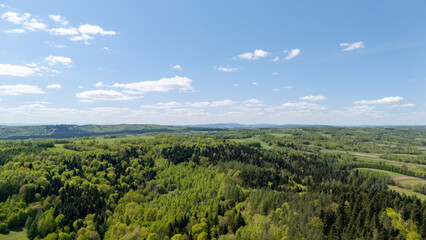 Aerial view of lush green forests and rolling hills under a blue sky with scattered clouds, showcasing the beauty of nature in springtime. Ideal for nature and travel themes.