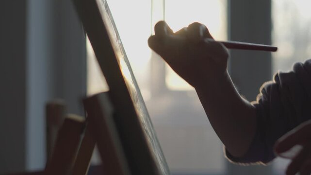 The image clearly illustrates a closeup view of a capable hand skillfully painting on a canvas bathed in warm sunlight, highlighting the peace, serenity, and passion inherent in the artistic process