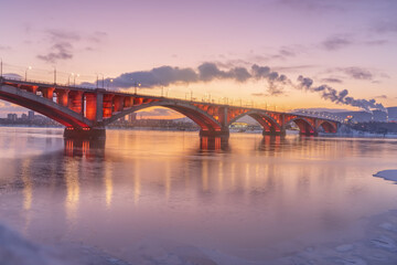 Naklejka premium Russia is an old bridge in the center of Krasnoyarsk across the Siberian Yenisei River, known as the Communal Bridge connecting the left and right banks of the city at the hour of colorful pink dawn.
