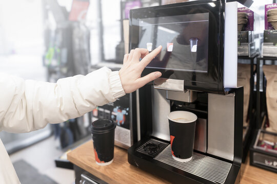 A woman pours and buys cappuccino coffee on her own at a self-service machine at a gas station or in a store. A close-up hand selects a drink with its finger on the touchscreen of a coffee machine.