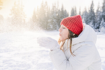 Young woman in winter attire playfully blowing snowflakes in a sunlit snowy landscape surrounded by evergreens
