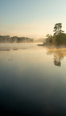 Fototapeta premium The tranquil lake reflects the soft light of dawn, with a gentle mist blanketing the waters surface and partially obscuring the distant shoreline, creating a serene and peaceful atmosphere at sunrise