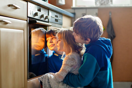 Little toddler girl and two kids boys baking pumpkin cake or cookies in kitchen. Three children, siblings, brothers and sister sitting near oven and waiting. On thanksgiving or christmas holiday. - Powered by Adobe