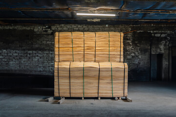 A large stack of plywood sheets tied with green straps, placed on pallets in a dimly lit industrial warehouse with exposed brick walls and ceiling.