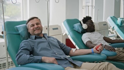 Portrait of happy middle-aged man in blue shirt lying in medical chair, donating his blood next to another donor in clinic